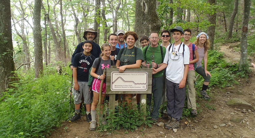 A group of parents and children pose for a photo near a sign in a green wooded area. The words on the sign are too small to read.